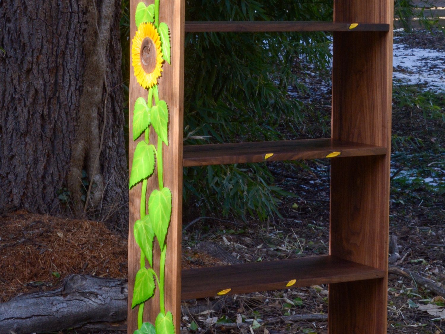 A solid walnut bookcase standing outdoors, open shelves on one side and sunflowers carved and painted by hand up the other