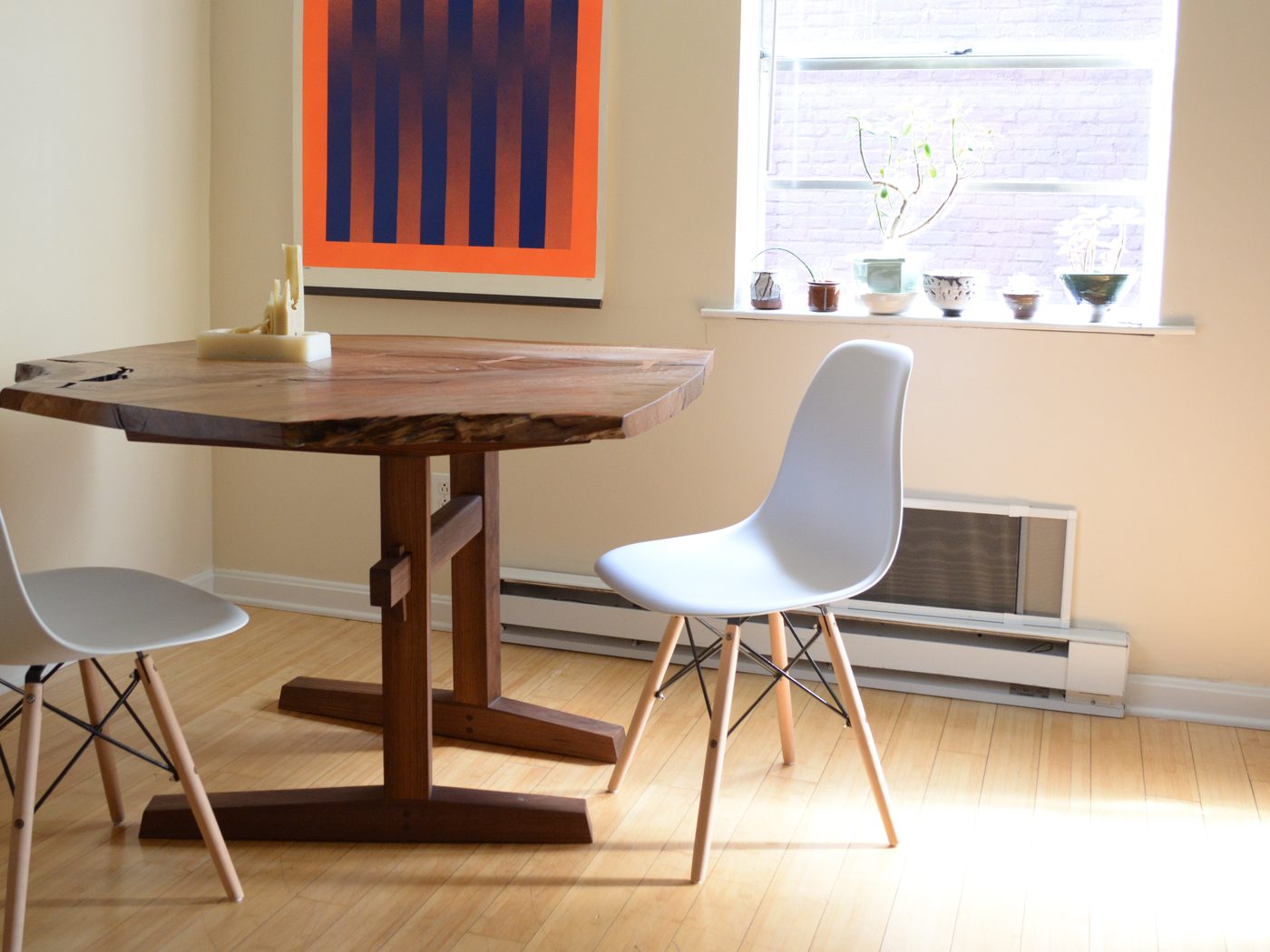 A live-edge dining table on a single central trestle whose feet splay outward, with pale chairs in a sunlit room